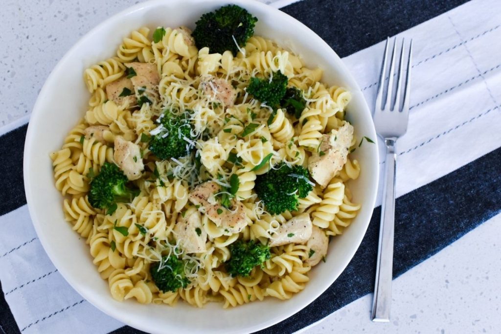 Rotini pasta, broccoli, chicken, parsley and parmesan cheese in a white bowl with a fork to the right of the bowl.