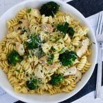 Rotini pasta, broccoli, chicken, parsley and parmesan cheese in a white bowl with a fork in the background.