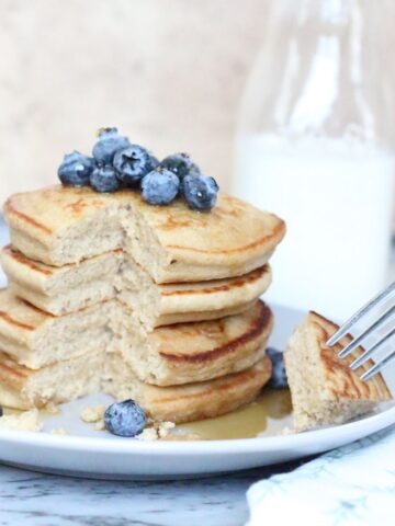 Stack tower of Oat Flour Pancakes on light gray plate, sliced to show fluffy texture, topped with blueberries. Milk is in glass behind plate of pancakes.