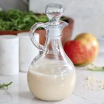 Apple Cider Vinaigrette dressing in glass container in front of red honey crisp apple, salt and pepper shakers, and a wooden bowl of greens.