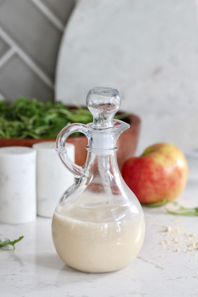 Apple Cider Vinaigrette dressing in glass container in front of red honey crisp apple, salt and pepper shakers, and a wooden bowl of greens.