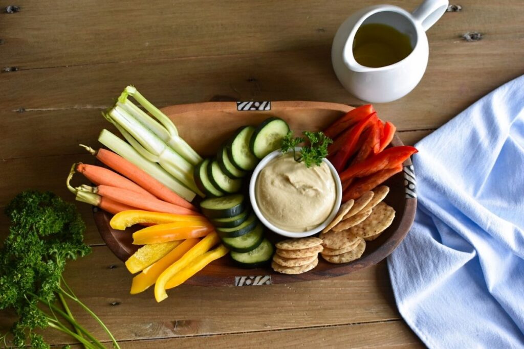 Hummus in a small dish with vegetables surrounding it in a wooden bowl and a blue kitchen towel and parsley on a the table.