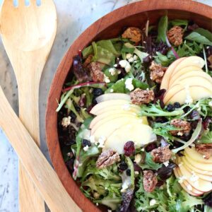 Apple Blue Cheese Salad in medium tone wooden bowl with light wood salad tongs. Salad has crumbles of blue cheese, candied pecans, dried cranberries, and fans of thinly sliced Honeycrisp apples on top.