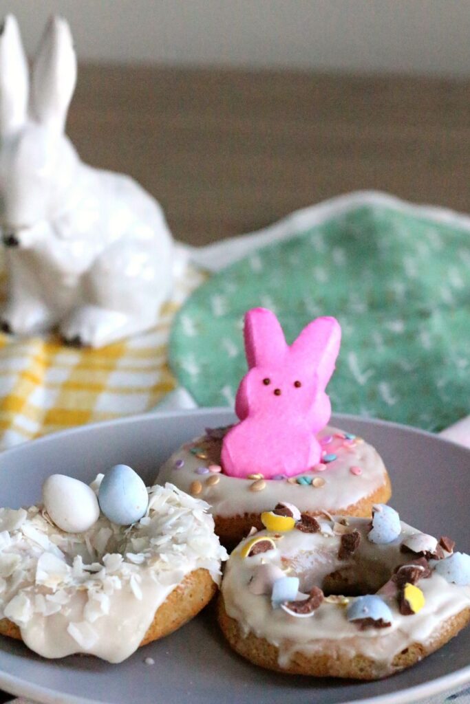 Three easter donuts: one with a pink marshmallow bunny, one with coconut shavings and mini Easter eggs and one with crushed chocolate candy coated easter eggs on top.