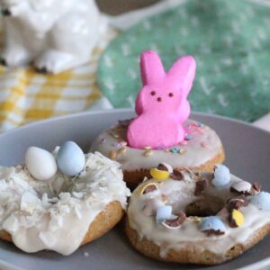 Three easter donuts: one with a pink marshmallow bunny, one with coconut shavings and mini Easter eggs and one with crushed chocolate candy coated easter eggs on top.