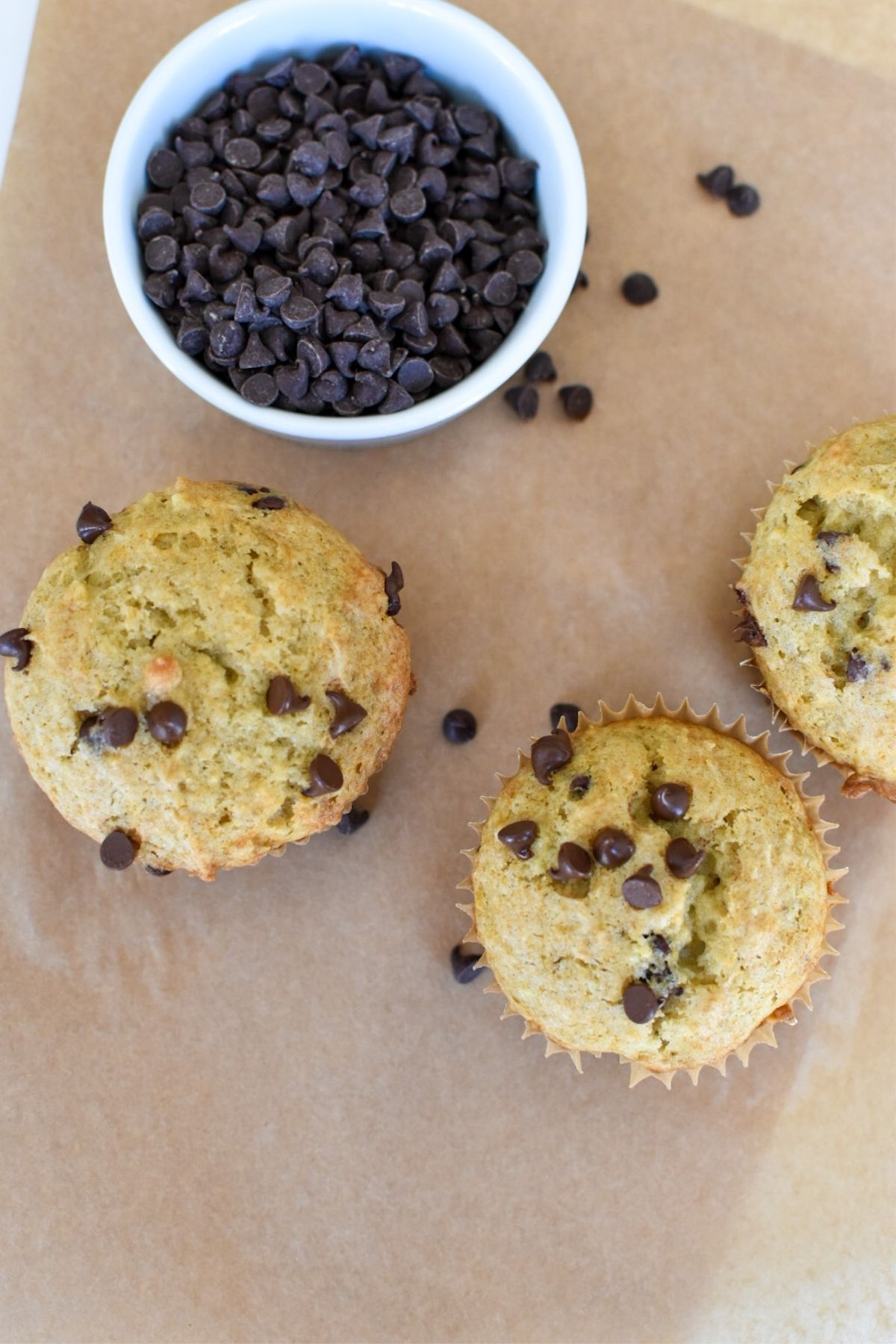 Three banana chocolate chip muffins with a small dish of mini chocolate chips in it.