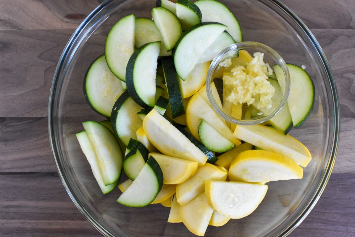 Zucchini and yellow squash cut into half circles in a glass bowl with minced garlic on top.