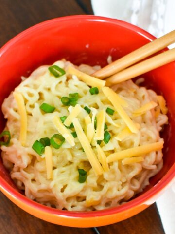 Cheesy ramen in a red bowl with cheese and green onions on top and chopsticks.