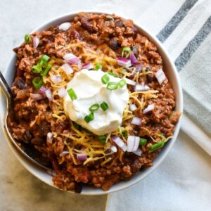 A bowl of chili with cheese, sour cream, green onions and red onions on top.