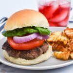 A burger with lettuce, onion and tomato next to french fries with a drink behind it.