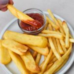 A french fry dipped in ketchup above a plate of steak fries and shoestring french fries.