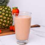 A smoothie in a glass with a strawberry on the rim and pineapple and strawberries in the background.