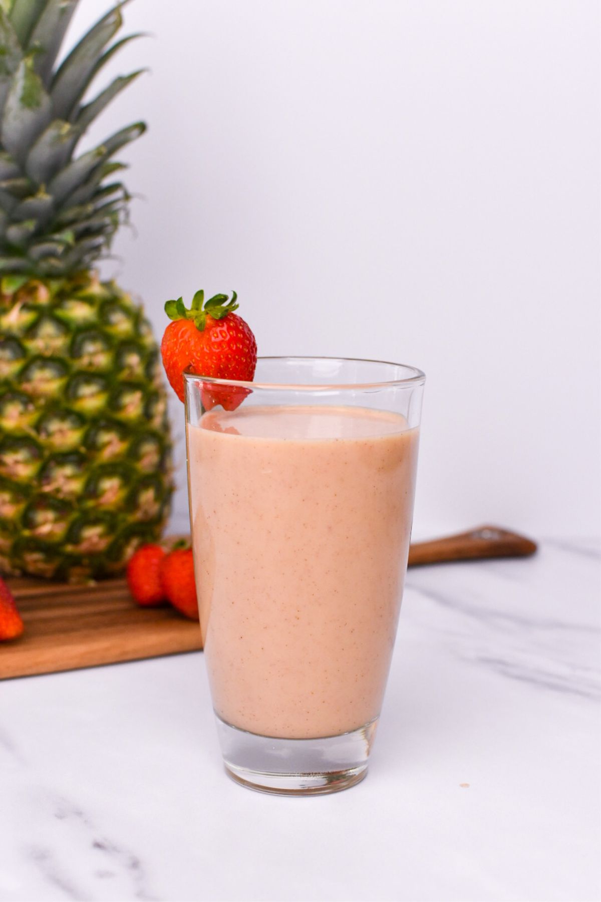 A smoothie in a glass with a strawberry on the rim and pineapple and strawberries in the background.