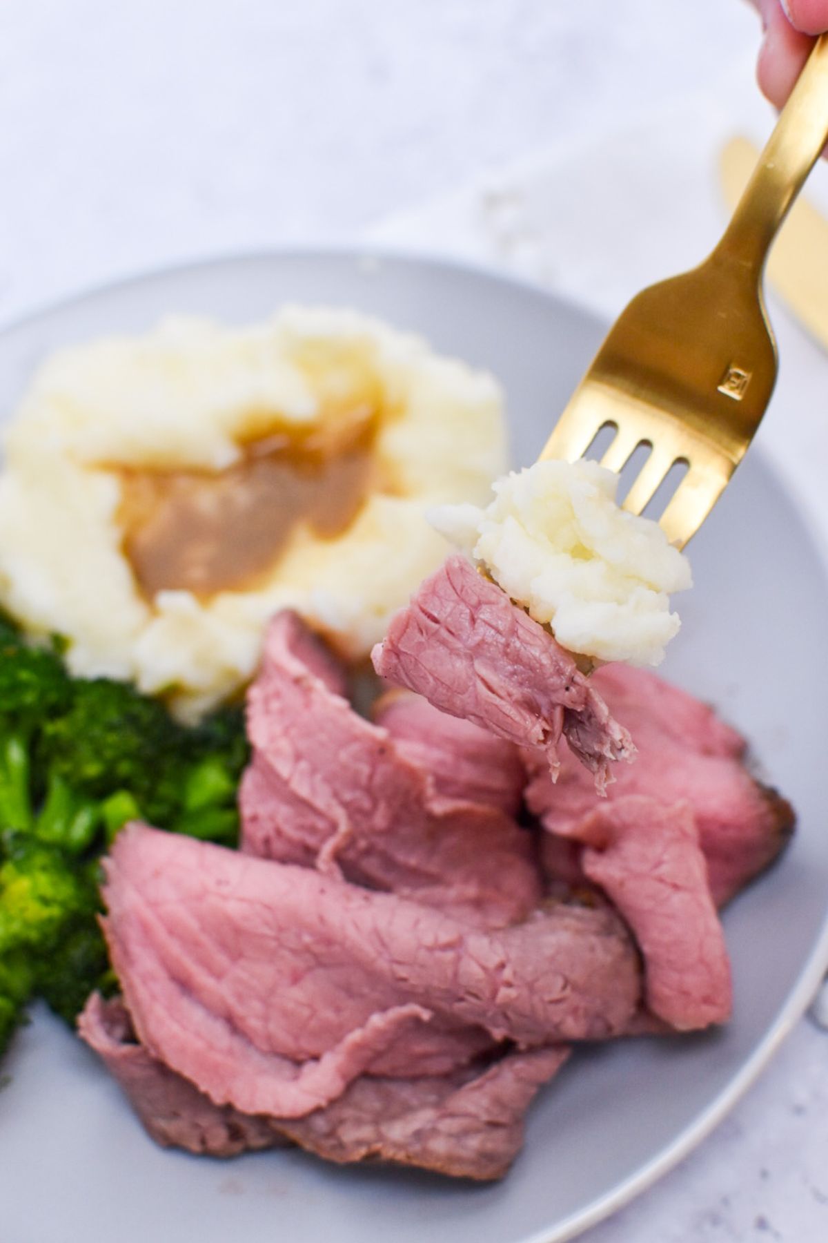 A gold fork with mashed potatoes and roast beef on it with a plate of food in the background.