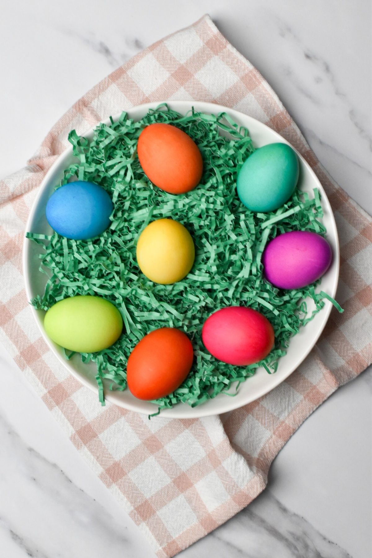 The colored eggs on green Easter grass in a white bowl with a white and pink kitchen towel under it.