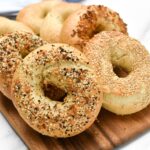 An everything bagel and sesame bagel with bagels stacked behind them.
