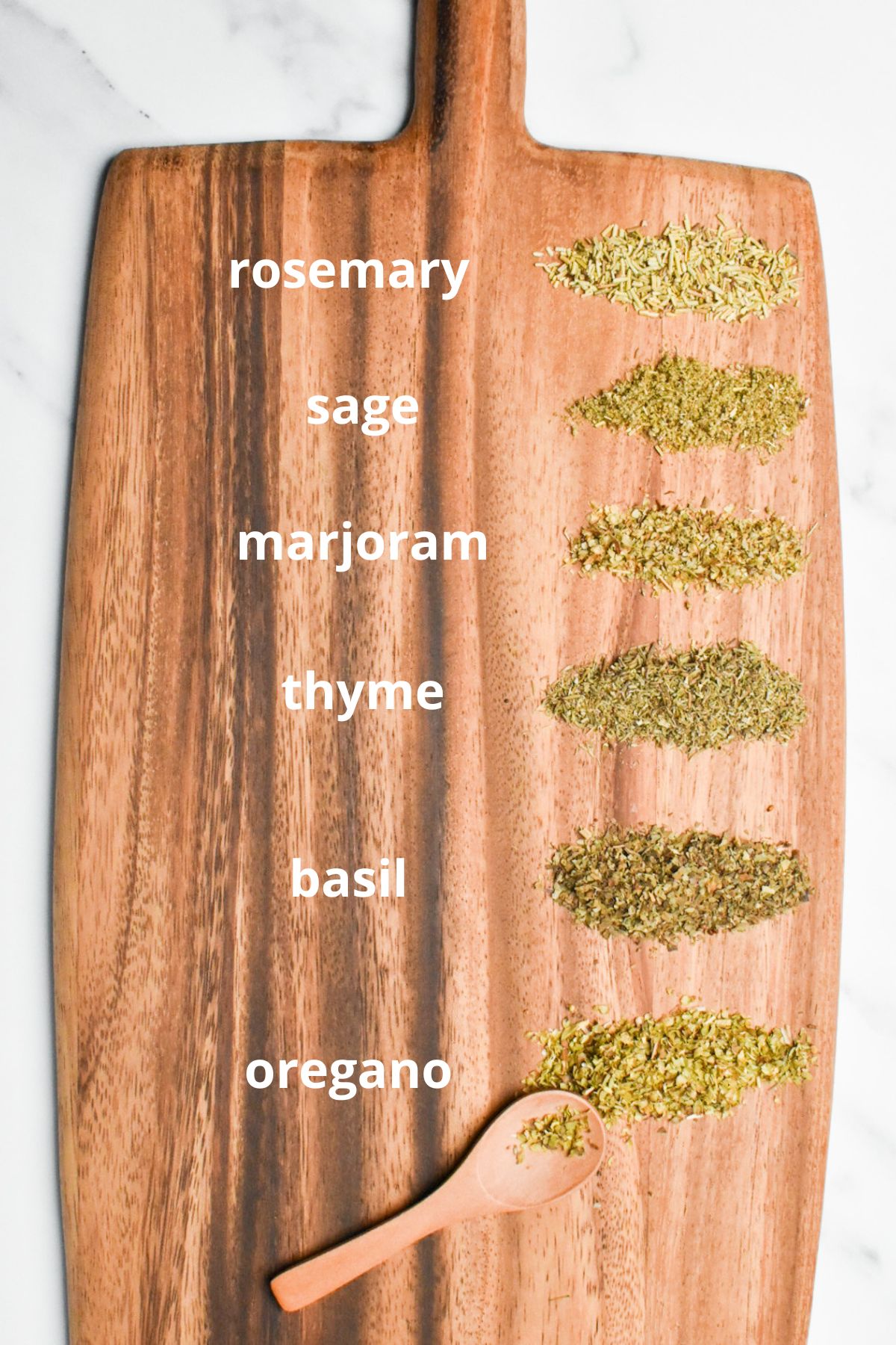 Dried herbs on a wooden board with white text next to them to label the rosemary, sage, marjoram, thyme, basil, and oregano.