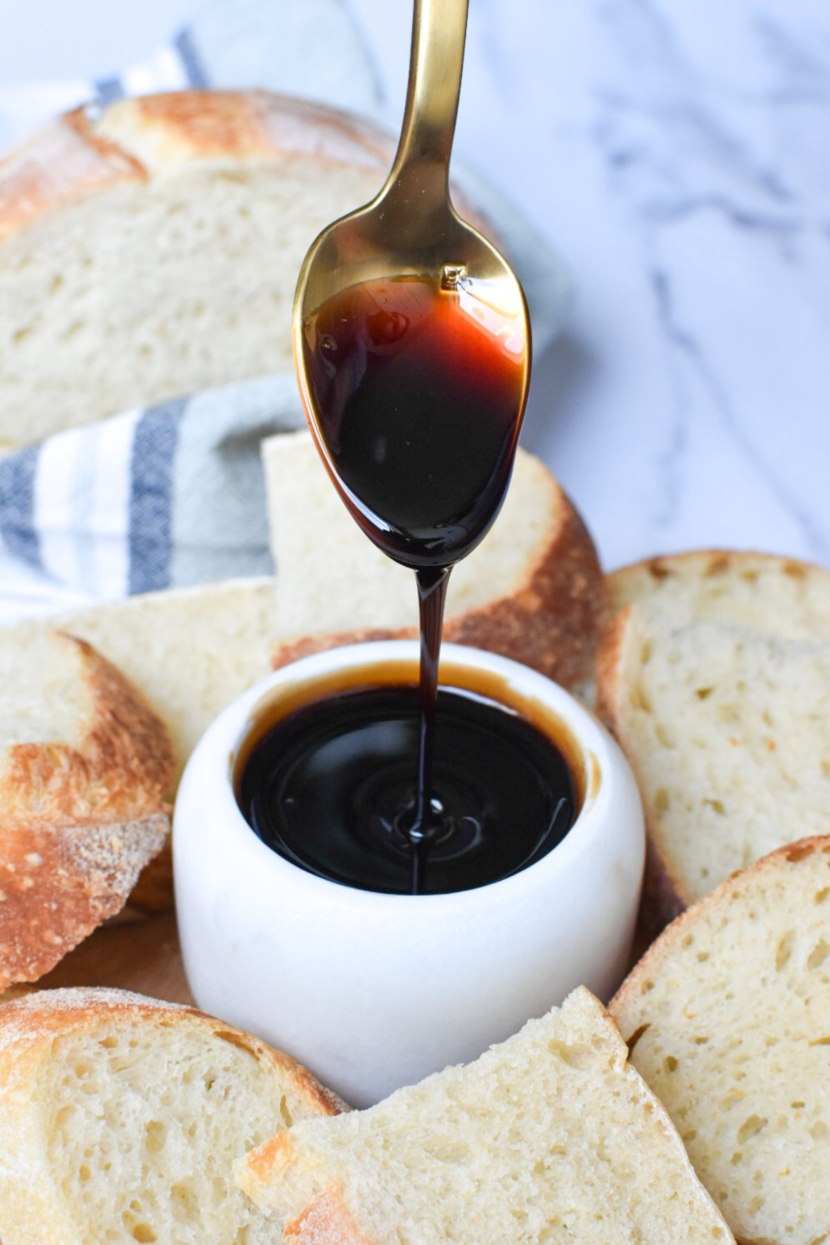 Thick caramelized balsamic vinegar being poured off a spoon into a small bowl with bread surrounding it.