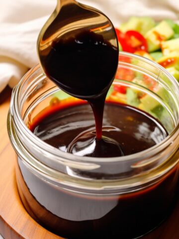 A small glass jar of balsamic glaze with the glaze being drizzled back into the jar from a spoon.