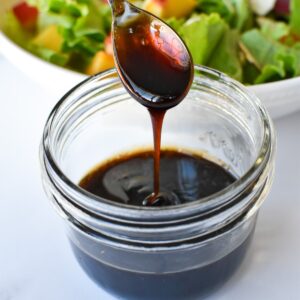 A small spoon drizzling balsamic glaze into a jar with a peach salad in the background.