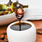 A small spoon drizzling thick balsamic fig glaze into a small white bowl.