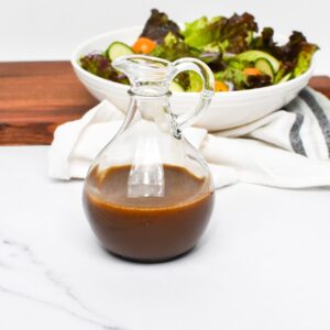 A glass salad dressing container with balsamic vinegar and olive oil dressing in it and a salad behind it.