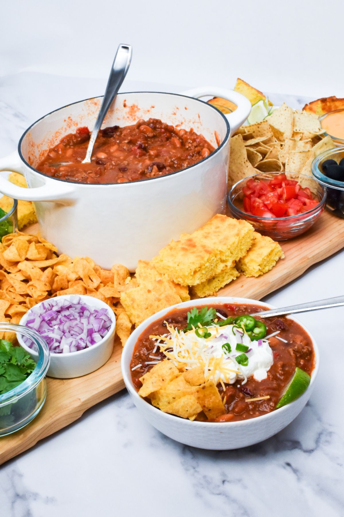 A bowl of chili and a wooden board full of toppings for chili.