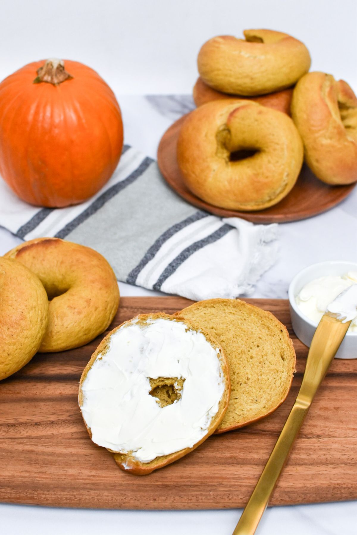 A sliced pumpkin bagel with cream cheese on a wooden board with bagels and a small pumpkin in the background.