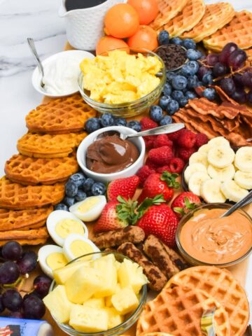 A long wooden board with waffles, pineapple, strawberries, syrup, sausage, eggs, bacon, blueberries, grapes, and more.