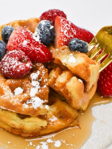 A fork taking a piece of sourdough waffles with fruit, powdered sugar, and syrup on top.