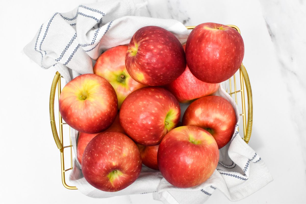 A gold basket with a kitchen towel in it and apples stacked inside.
