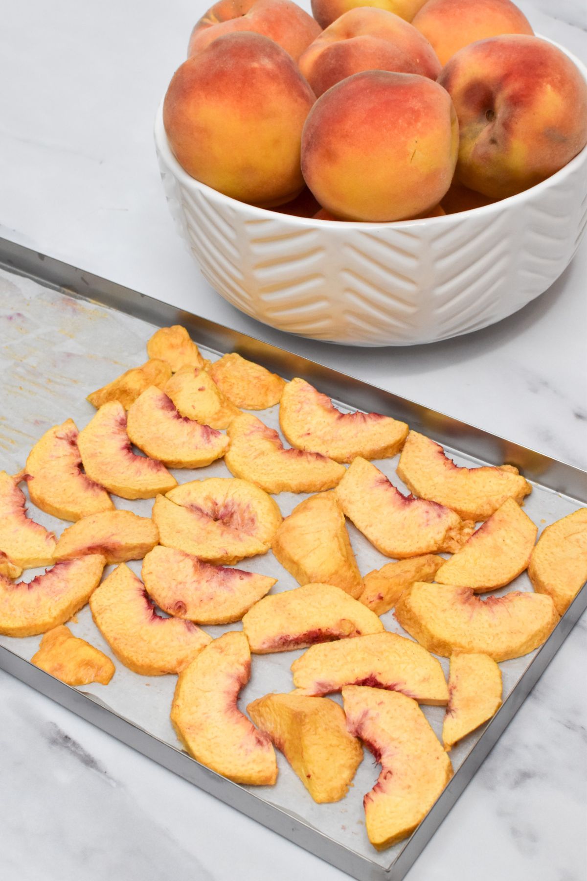 A metal tray of freeze dried peaches on parchment paper with a white bowl of peaches behind it.