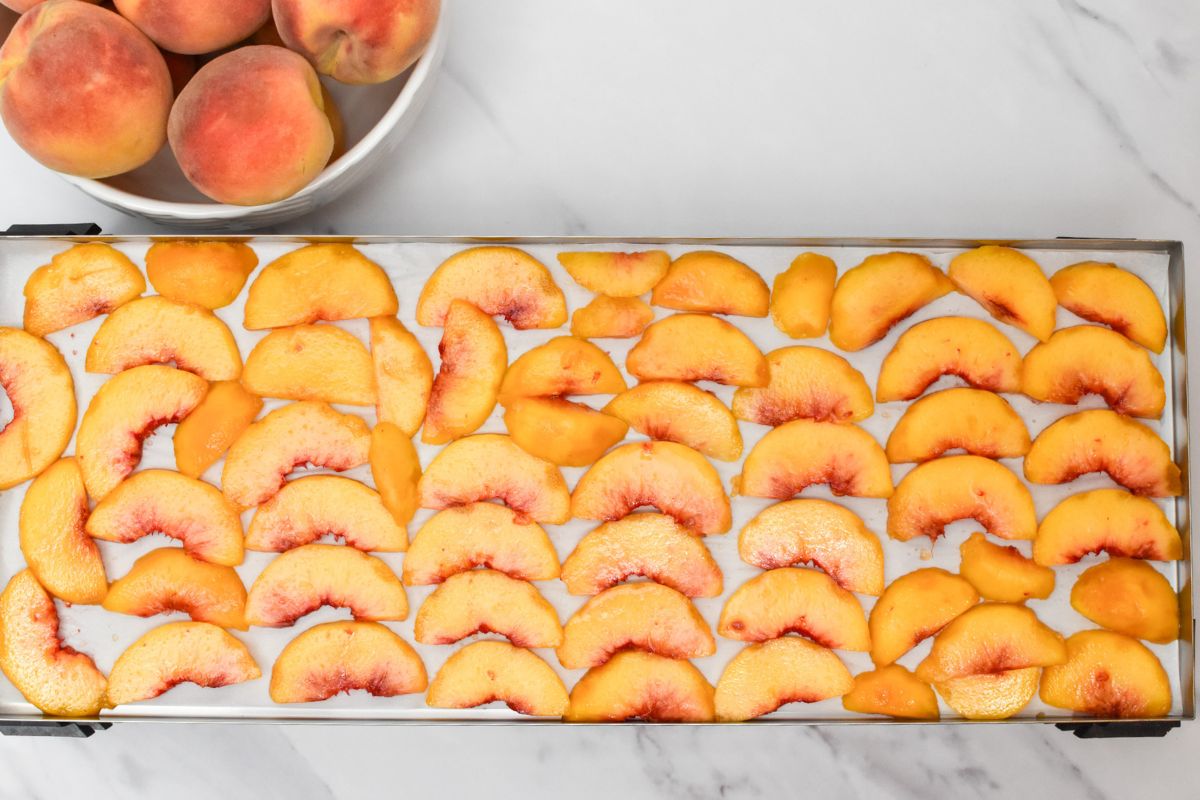 A metal tray of sliced peaches with a bowl of peaches behind it.