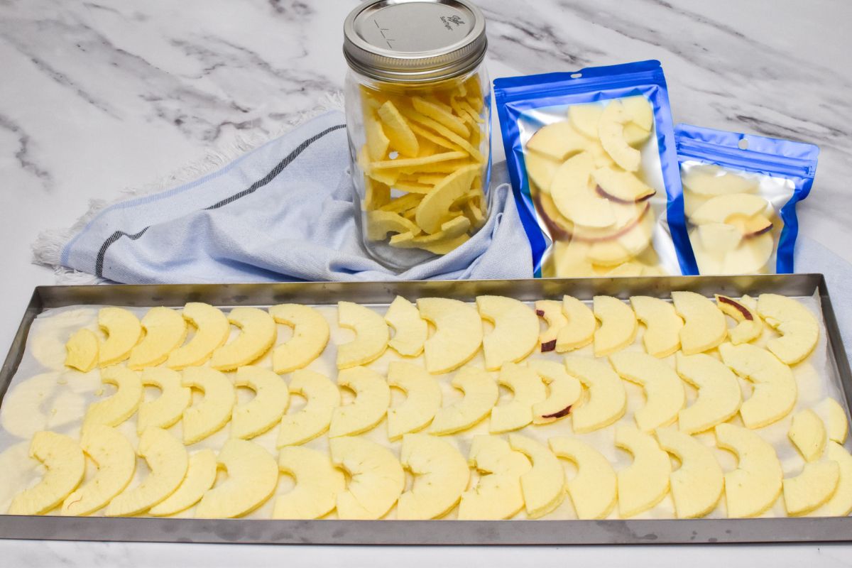 Freeze dried apples on a metal tray with a mason jar and mylar bags of freeze dried apples behind the tray.