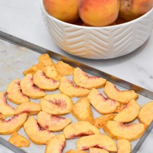 A metal tray of freeze dried peaches on parchment paper with a white bowl of peaches behind it.