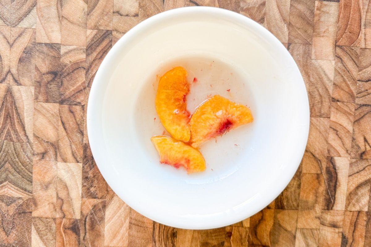 A white bowl with water and 3 slices of peaches in it on a wooden cutting board.