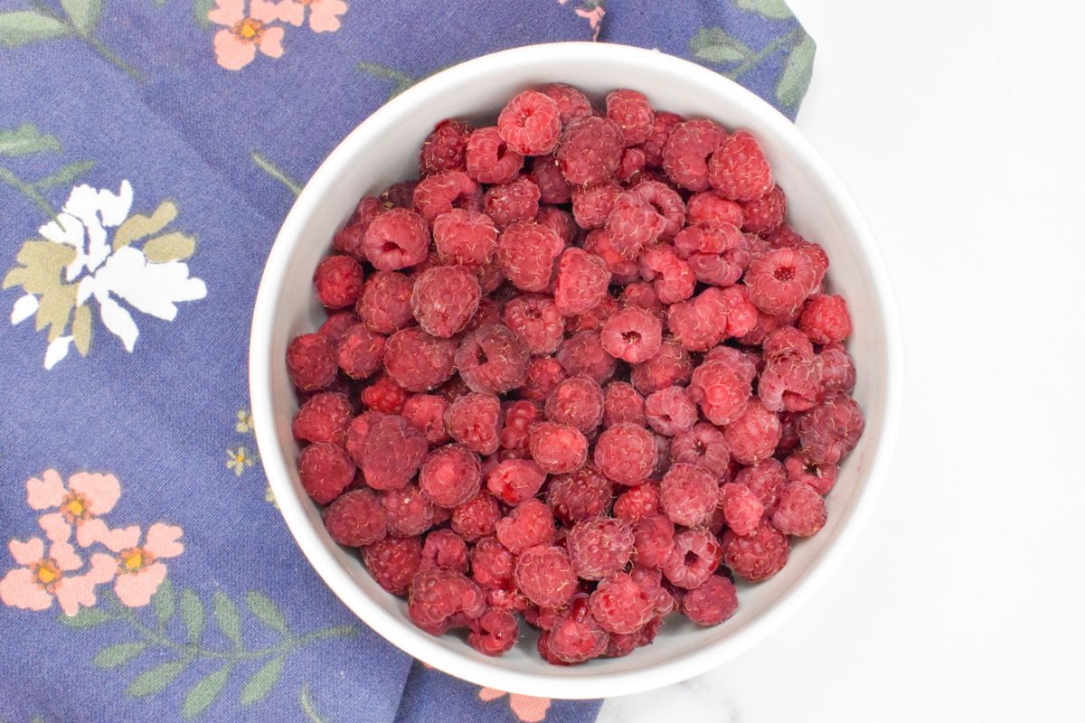 A bowl of fresh raspberries on a towel with flowers printed on it.