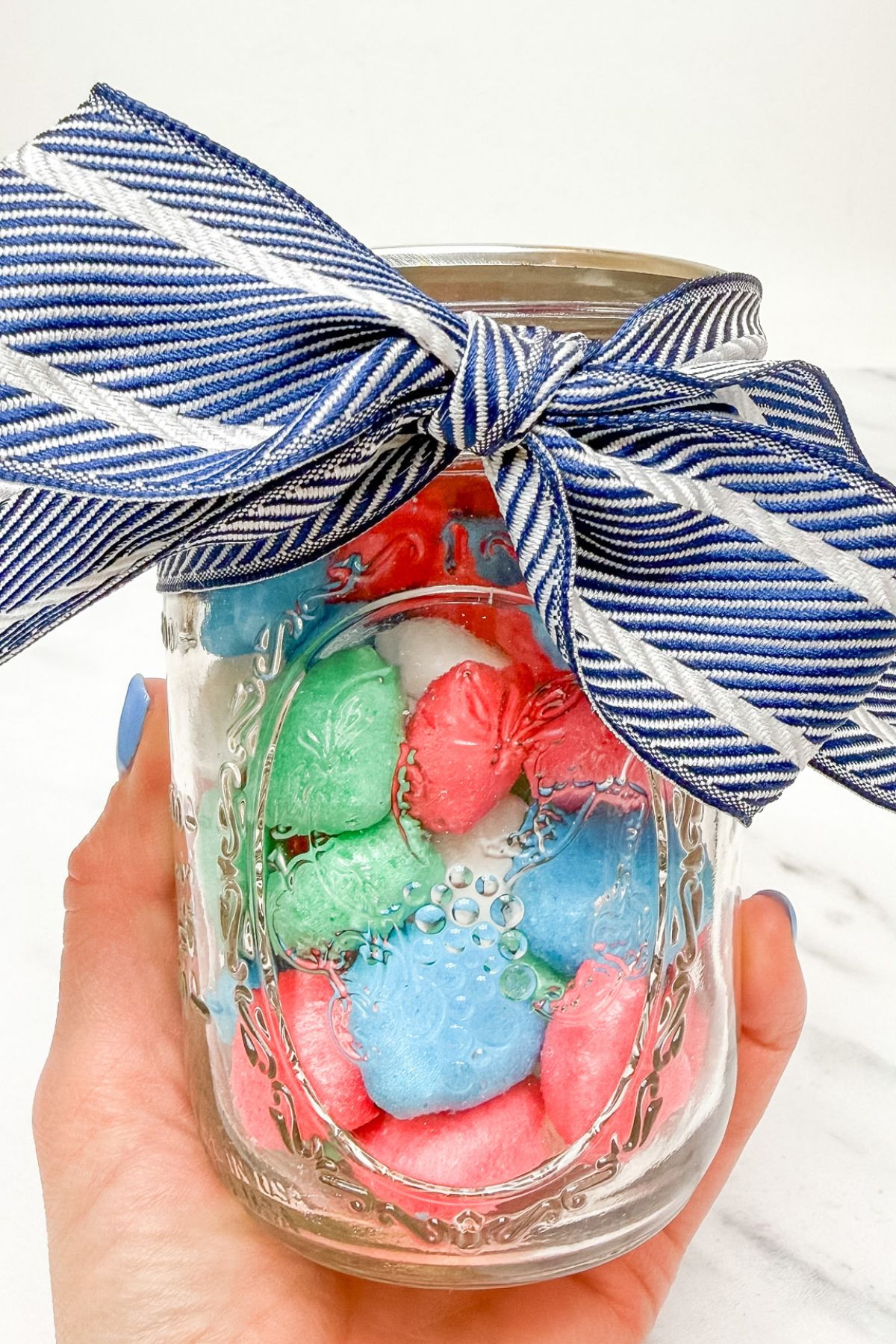 A hand holding a glass jar of freeze dried Airheads with a blue ribbon tied to the top of the jar.